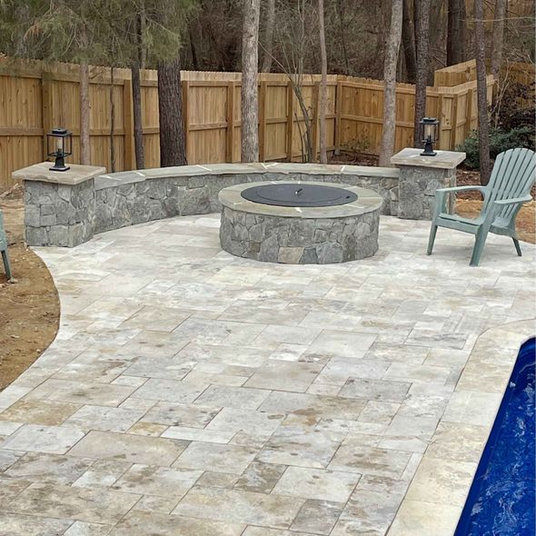 Stone patio with curved retaining wall, fire pit, and poolside chair beside a fenced yard