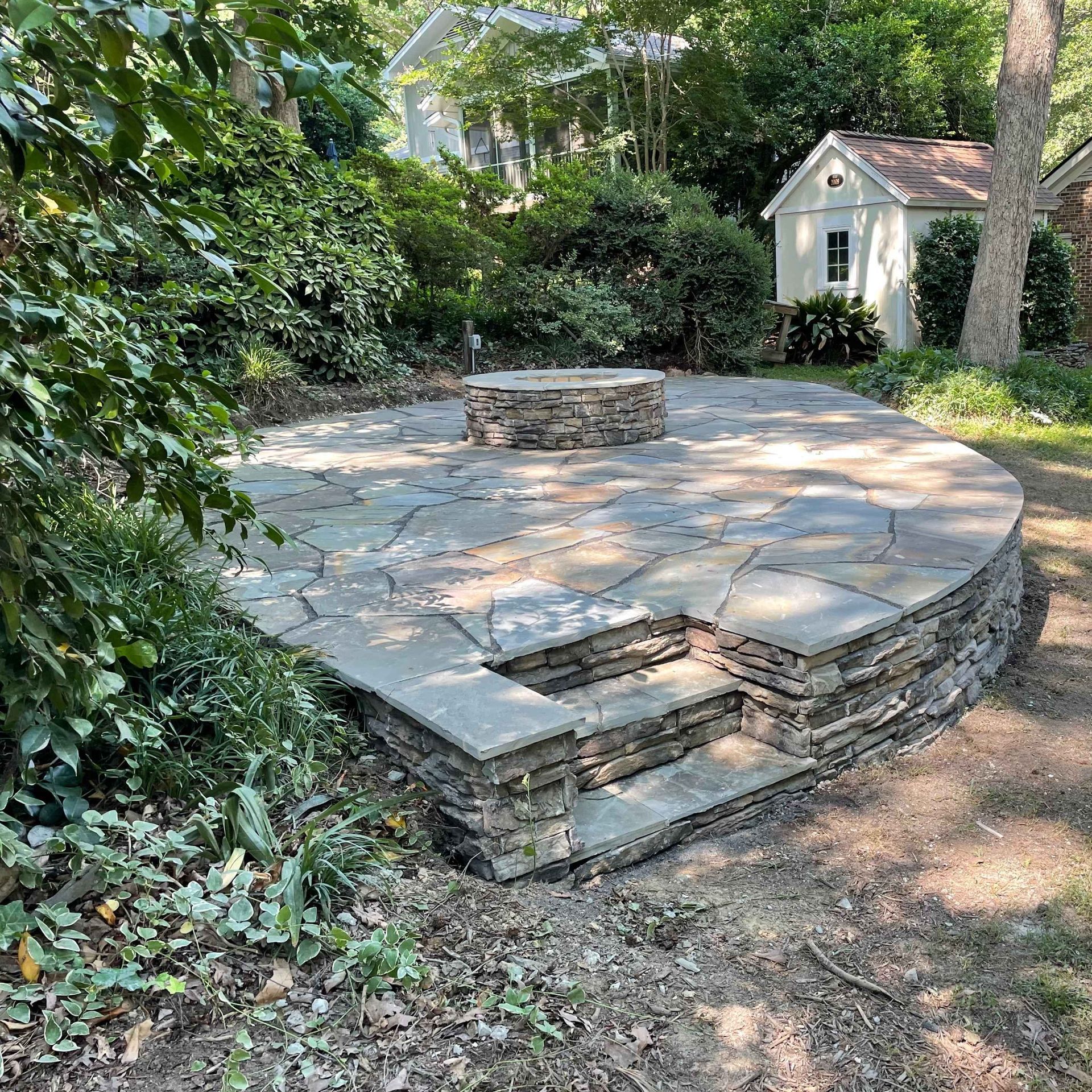 Circular stone patio with central fire pit and steps, surrounded by lush greenery and a small shed