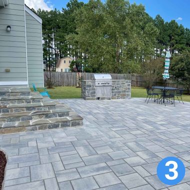 Stone patio with built-in grill, steps by a house, and outdoor table near a fenced backyard with trees.