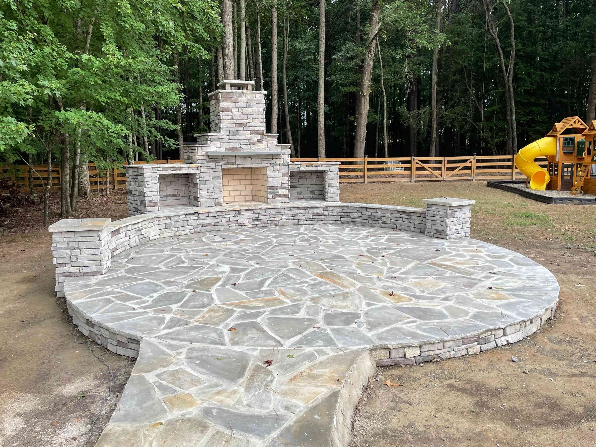 Circular stone patio with a white stone monument, surrounded by trees and a wooden fence at a worksite