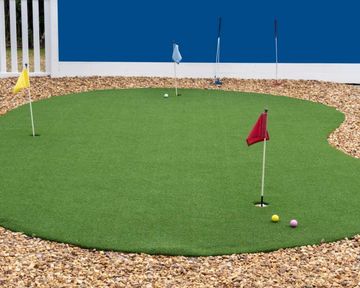 Small artificial putting green with colored flags and golf balls beside a blue wall and white fence.