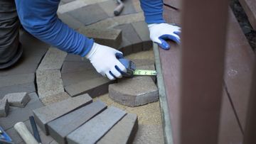 Gloved hands measuring gaps between gray paving stones during patio repair