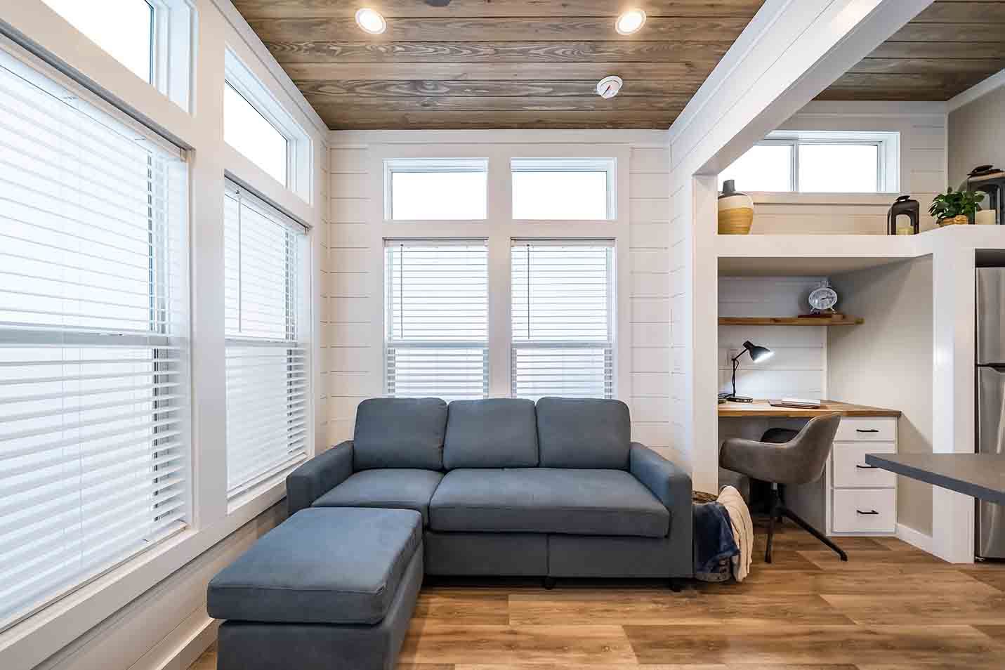 Bright modern living room with gray sectional, wood ceiling, large windows, and desk nook