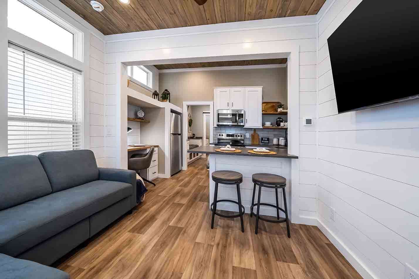 Small modern studio apartment with gray sofa, two stools, and compact kitchen with wood floors and white walls