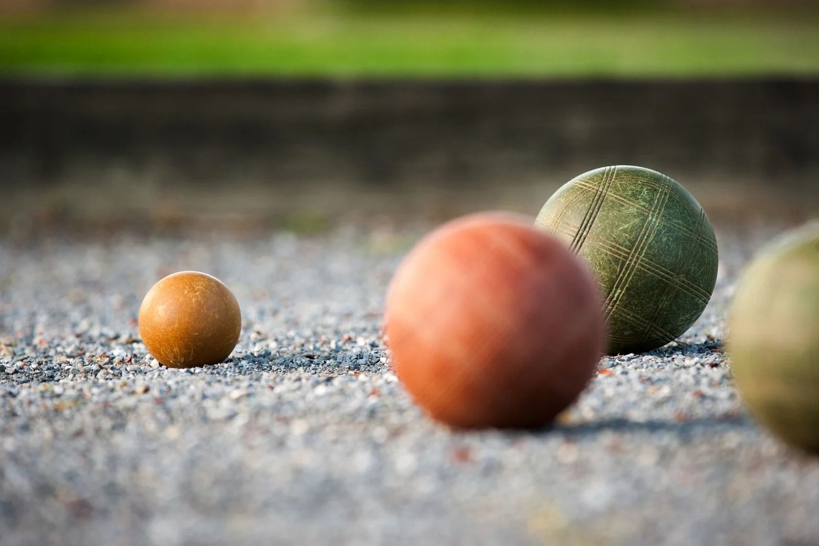 Three bocce balls on a gravel court, with orange, green, and pale yellow balls in shallow focus.