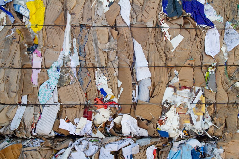 Torn paper and cardboard scraps in beige, blue, white, and yellow, stacked in an industrial recycling bale.