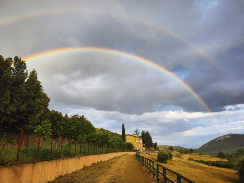 Un doppio arcobaleno si inarca su un sentiero che si snoda attraverso un paesaggio con un campo, alberi e un edificio.