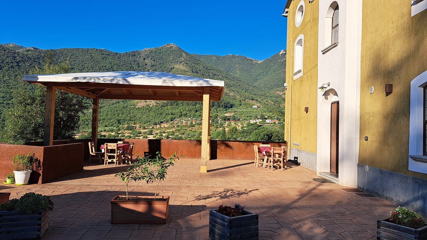 Edificio giallo con terrazza ombreggiata con vista sulle montagne. Tavoli e sedie sul patio.