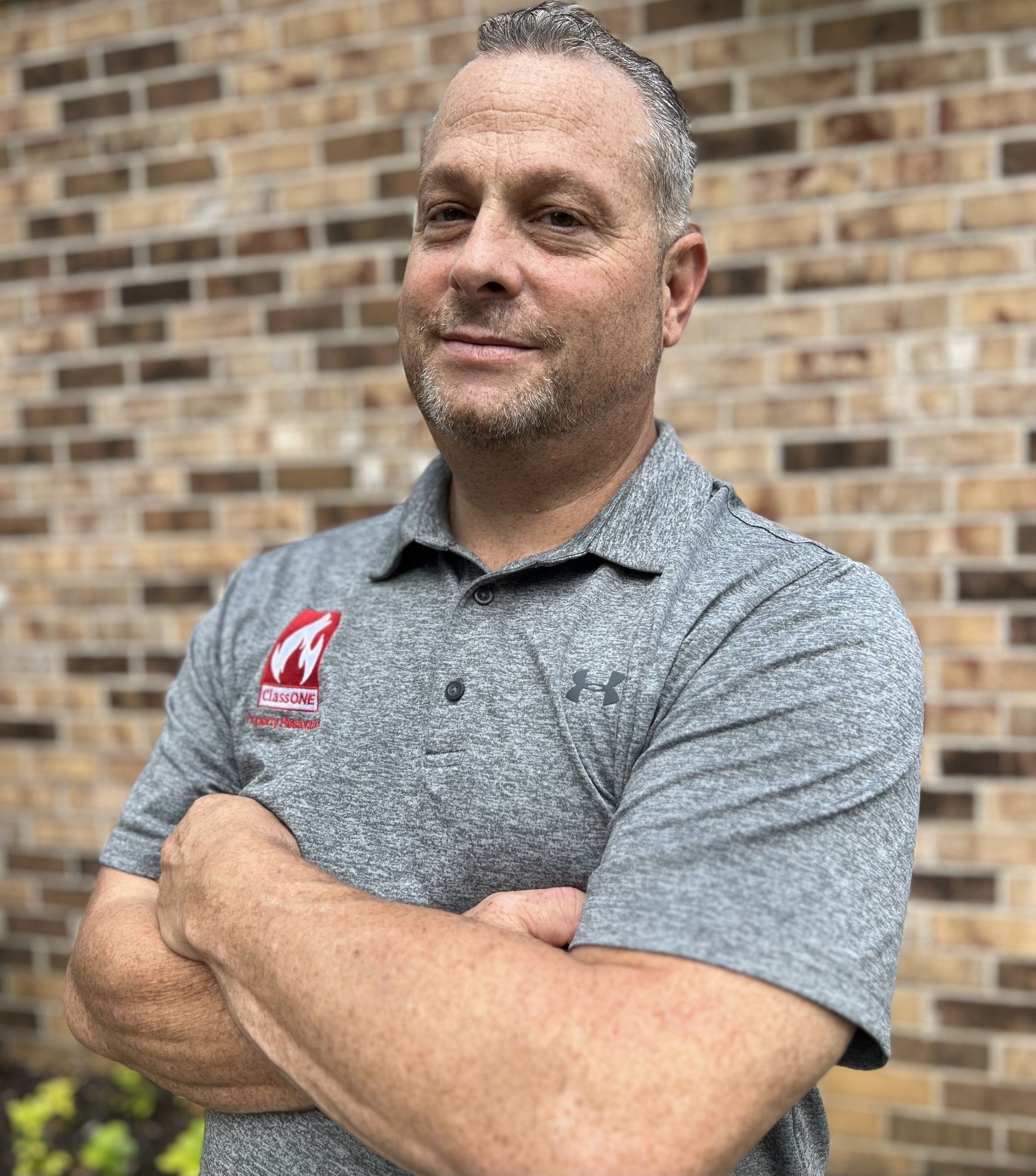 Man with arms crossed, wearing gray polo shirt with logo, standing in front of brick wall.