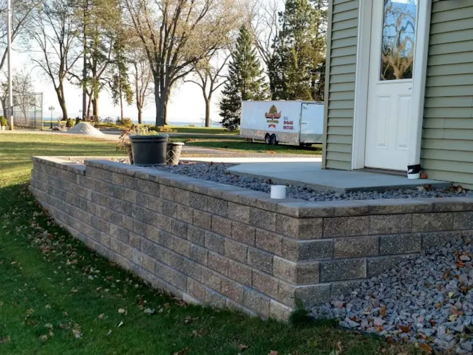 Brick patio beside a house with a white door, a black planter, and a yard with trees in the background.