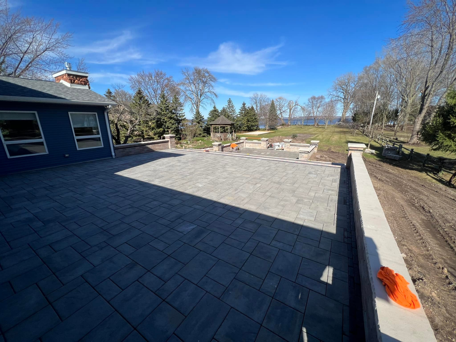 Large stone patio beside a blue house, with a clear sky, trees, and a landscaped yard beyond.