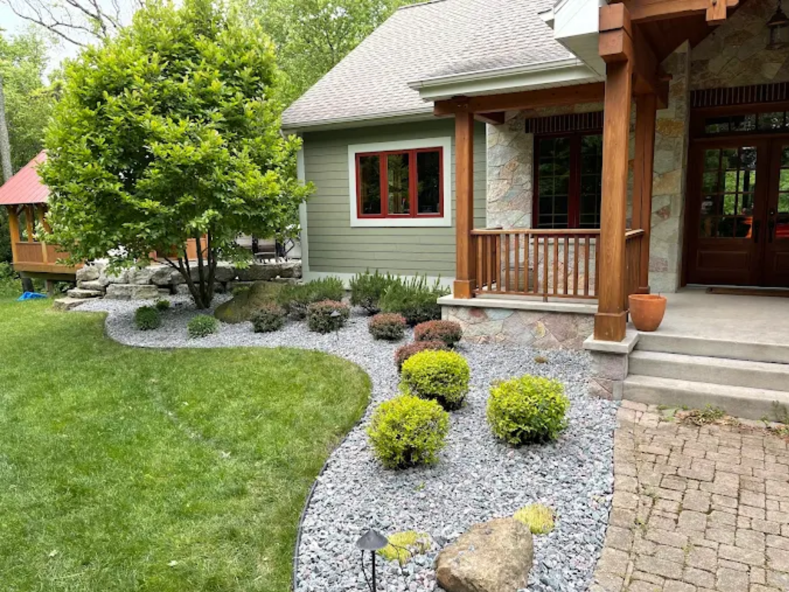 Two men shake hands in a sunny suburban yard beside a flower bed and house.