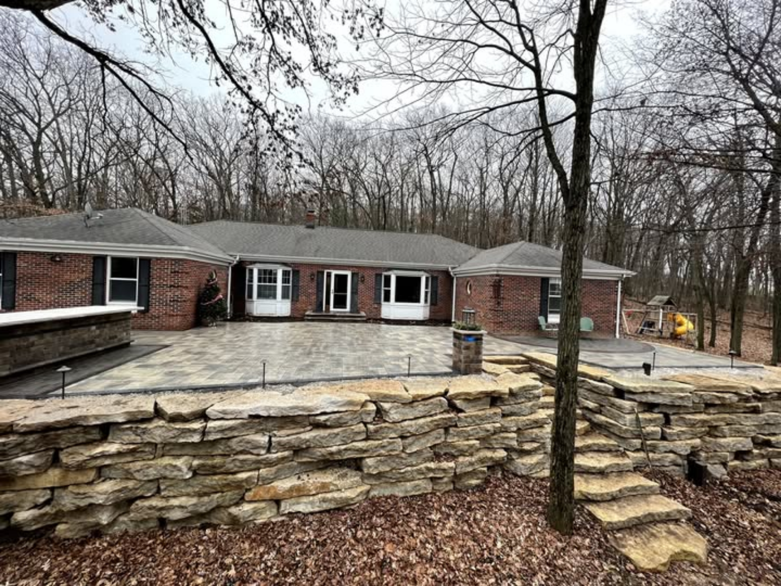 Brick house with a large stone patio and steps, surrounded by leafless trees in a wooded yard