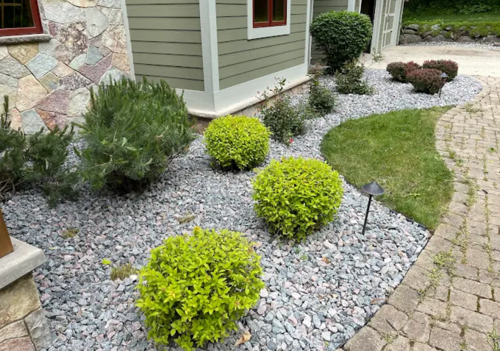 Front yard garden with gray gravel, small green shrubs, and a curved brick walkway beside a house.