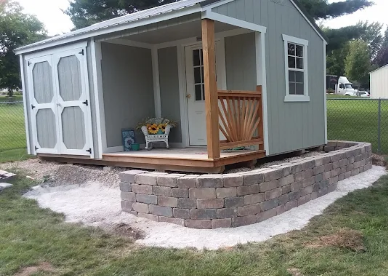Small gray shed with porch and double doors, sitting on a raised stone retaining wall in a grassy yard