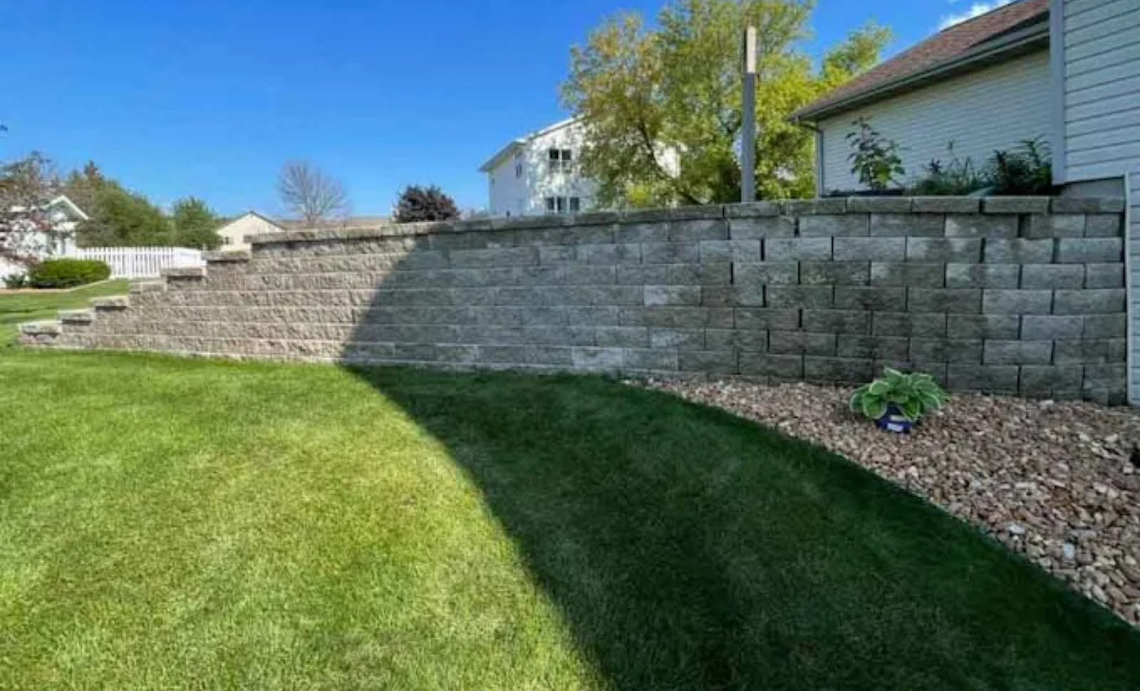Curved green lawn beside a gray stone retaining wall and white house under a clear blue sky