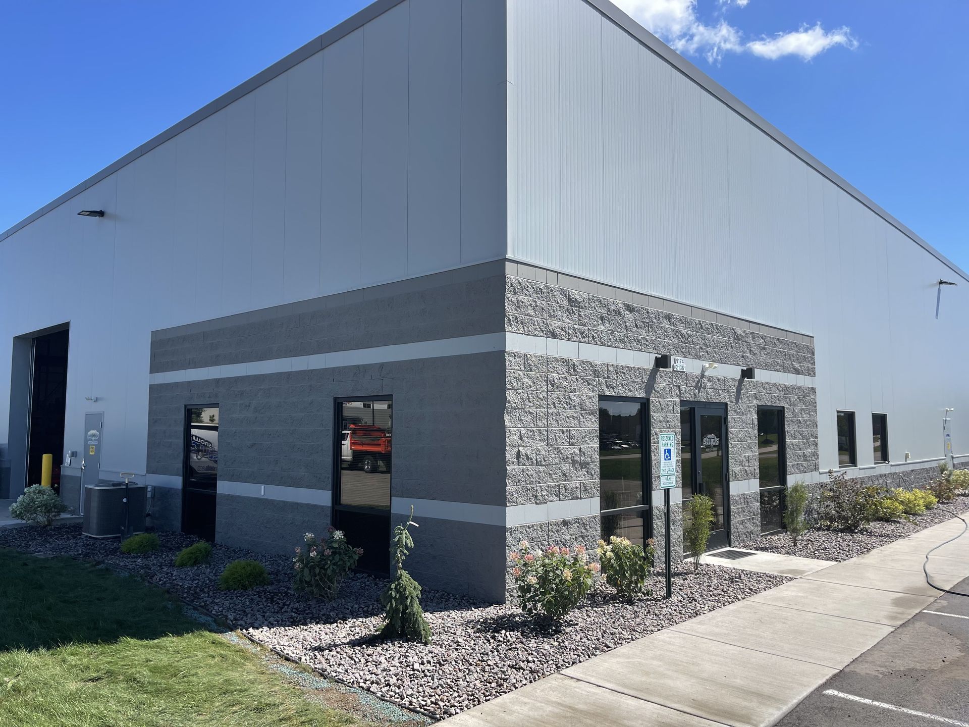 Industrial building exterior with gray metal siding, stone accents, and a sidewalk under a blue sky