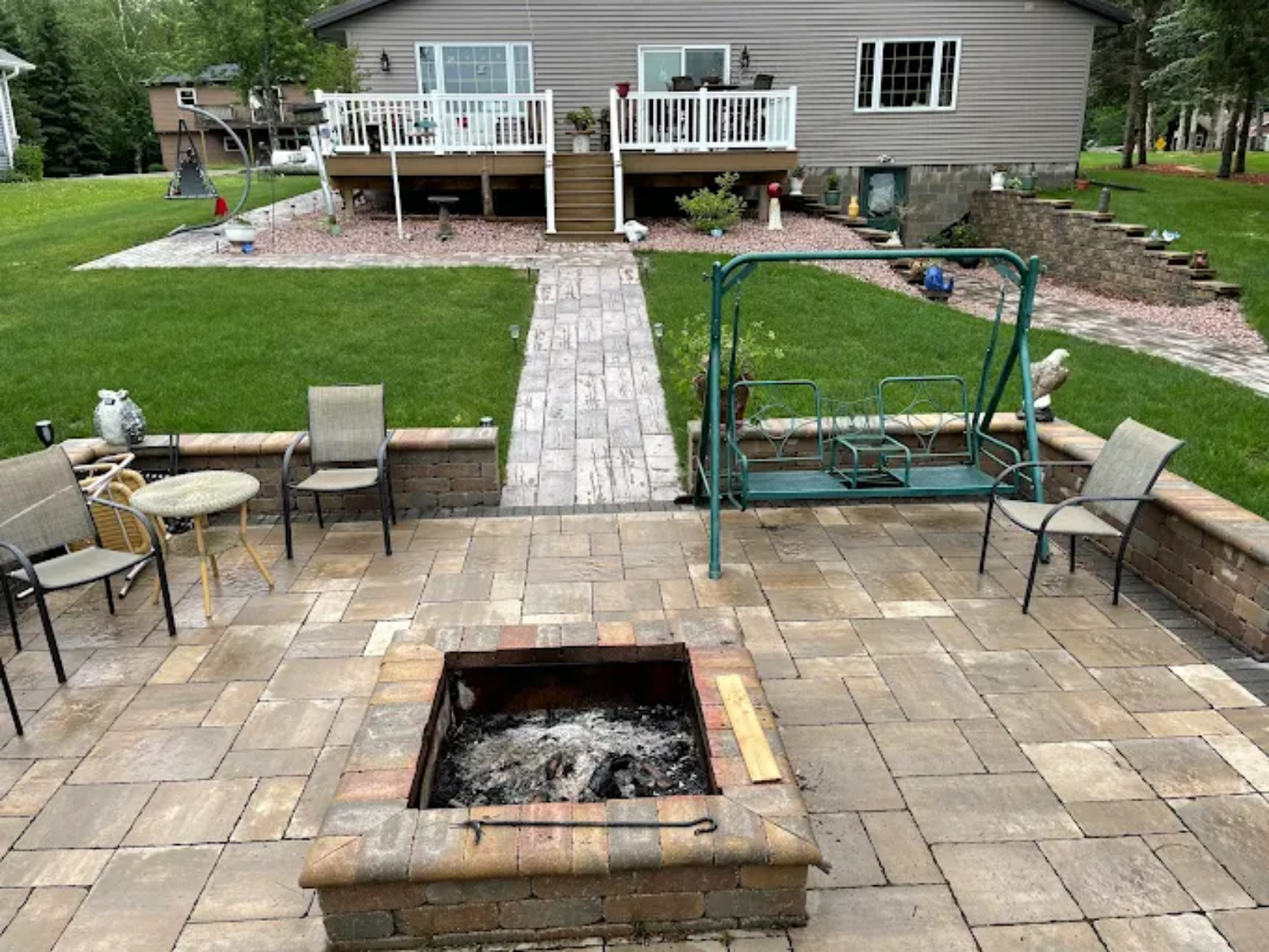 Backyard patio with fire pit, chairs, swing, lawn, and deck stairs leading to a house.
