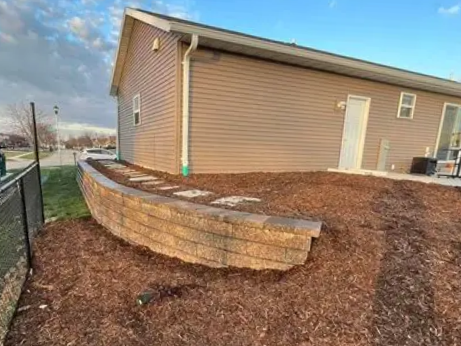 Curved stone retaining wall beside a tan house, with fresh mulch on a sloped yard.