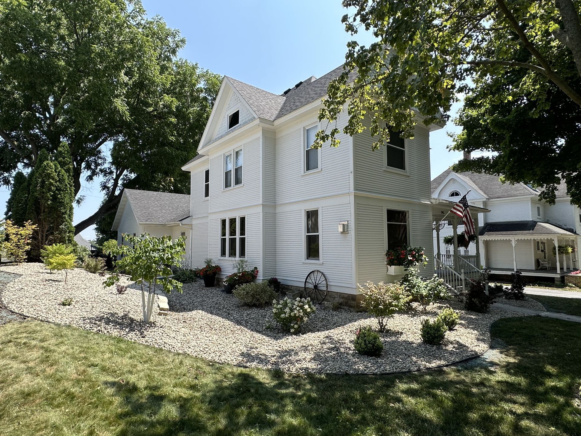 White two-story house with gray roof and stone landscaping, viewed from a leafy front yard