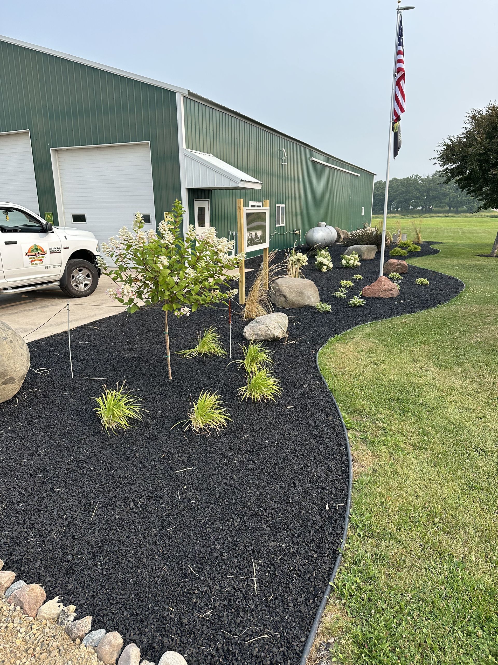 Green building with black mulch landscaping, white pickup truck, and U.S. flag on a grassy roadside.