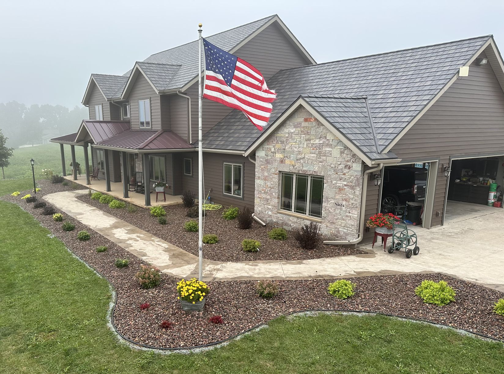 Stone house with an American flag, flower beds, and a driveway under a cloudy sky