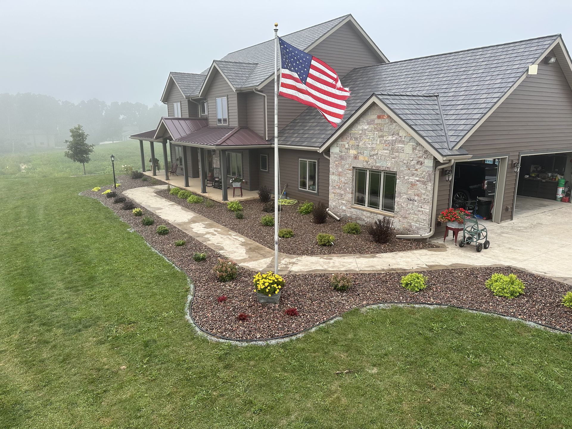Stone house with porch and large American flag beside a landscaped lawn on a misty day