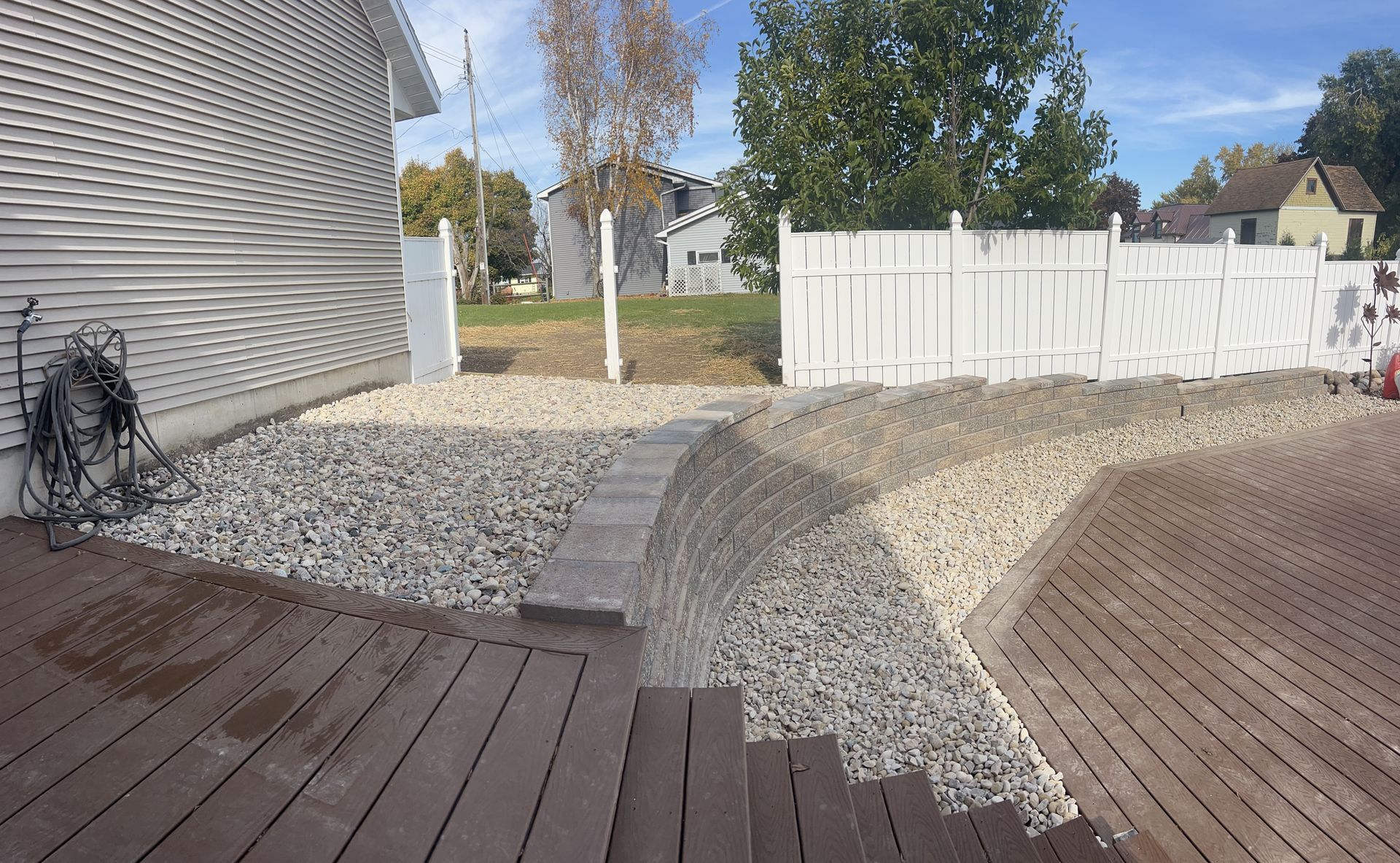 Backyard patio with gravel path, white fence, and trees on a sunny day