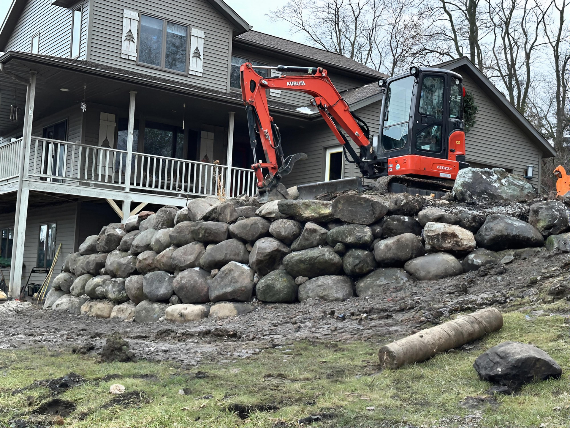 Orange excavator on a large rock retaining wall beside a gray house and porch outdoors