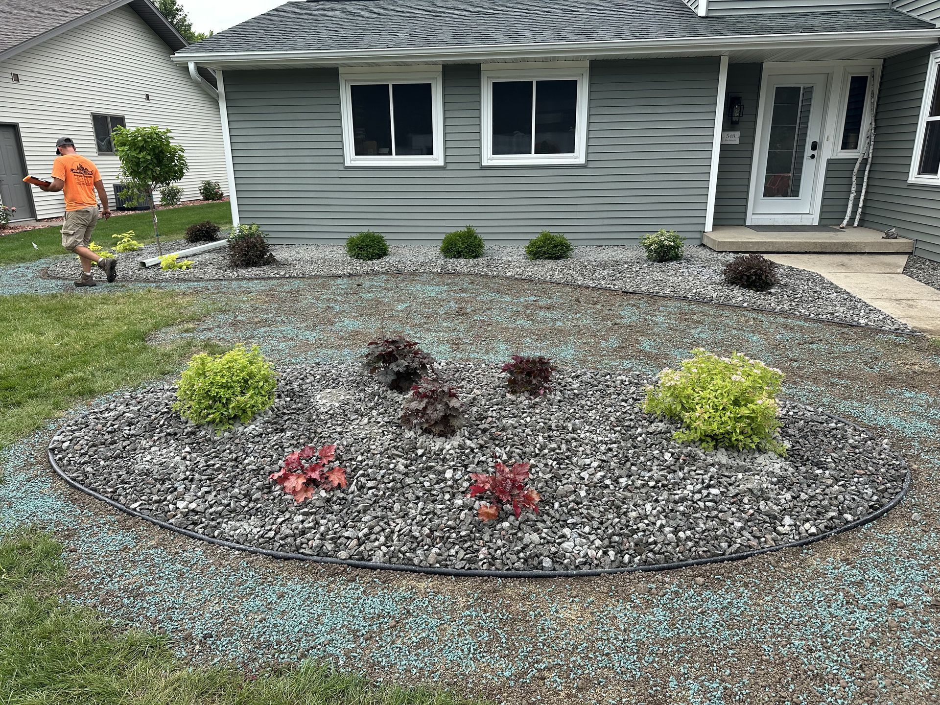Front yard landscape with mulch bed, small shrubs, and a gray house with windows and a front door.