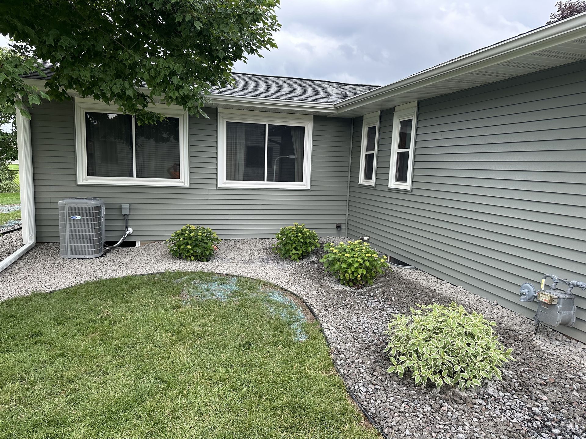 Small gray-sided house courtyard with curved gravel bed, green lawn, shrubs, and window air conditioner