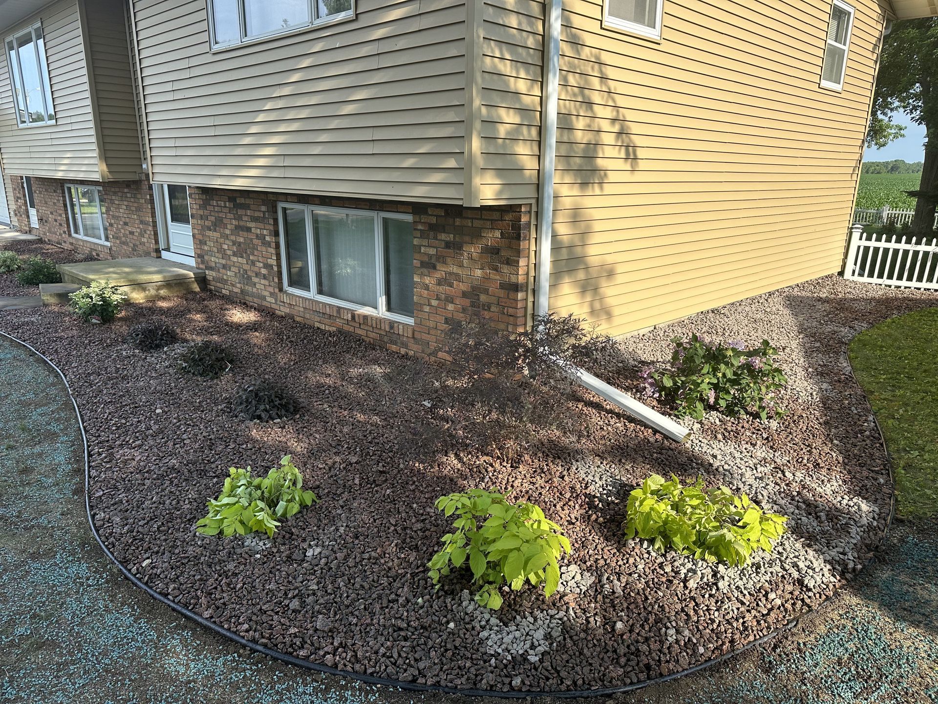 Mulched landscaping beds with small shrubs along the side of a tan house with brick and siding