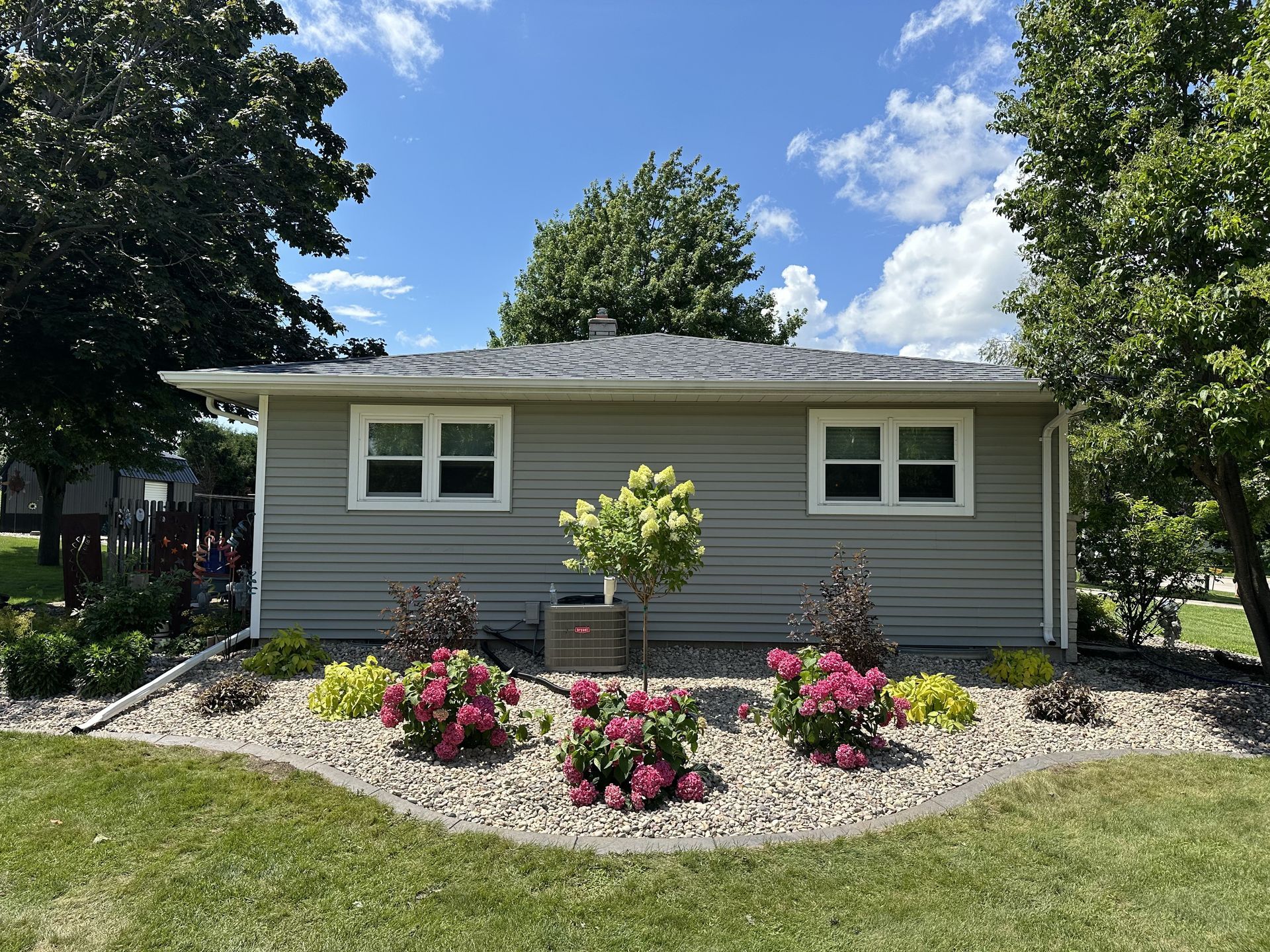 Small gray house with white trim, flower beds, and a green lawn under a blue sky
