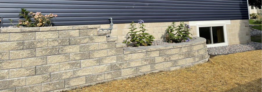 Stone retaining wall with small shrubs in front of a dark-sided house and basement window
