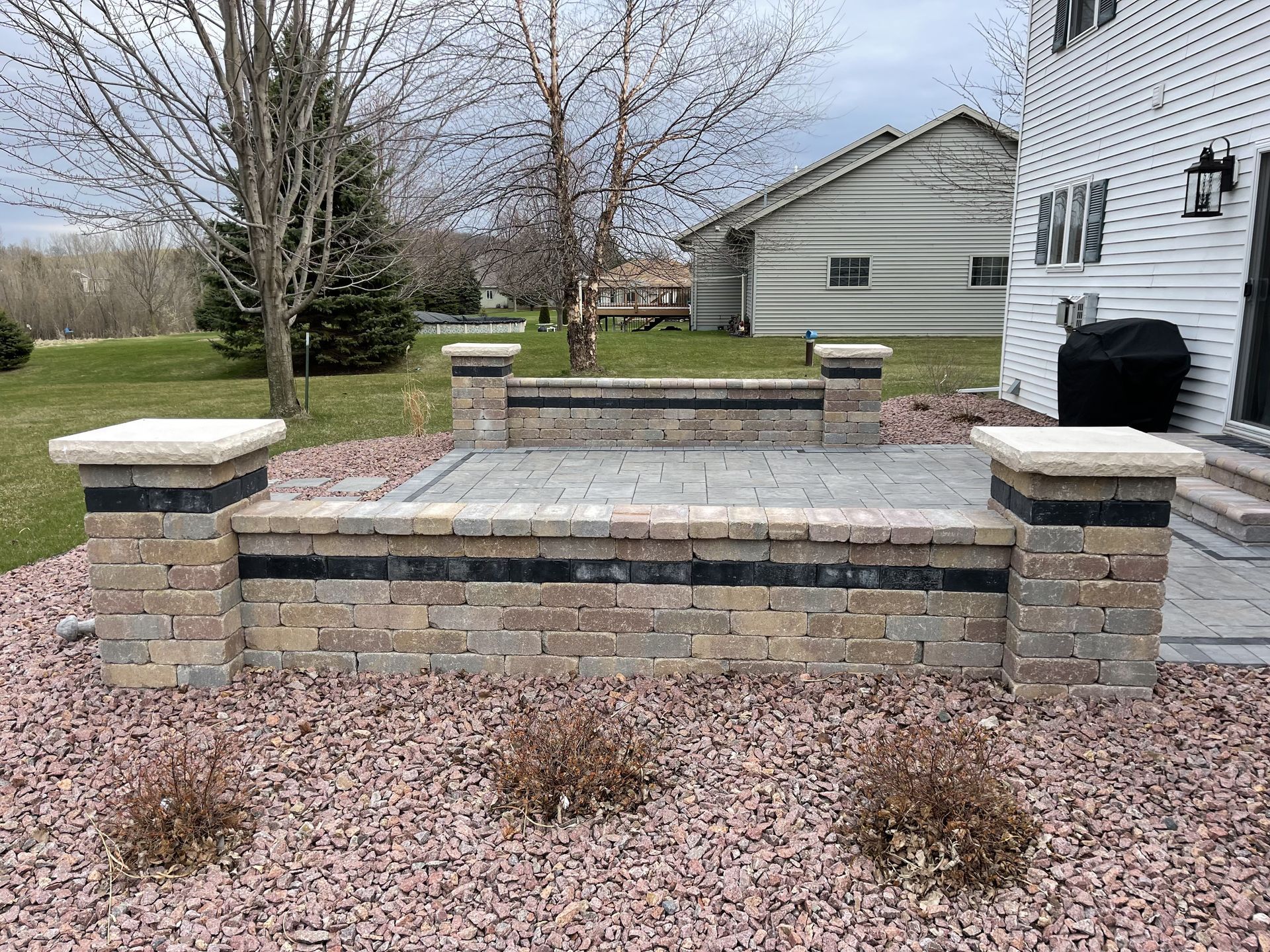 Brick raised patio with stone pillars and steps in a backyard beside a white house.