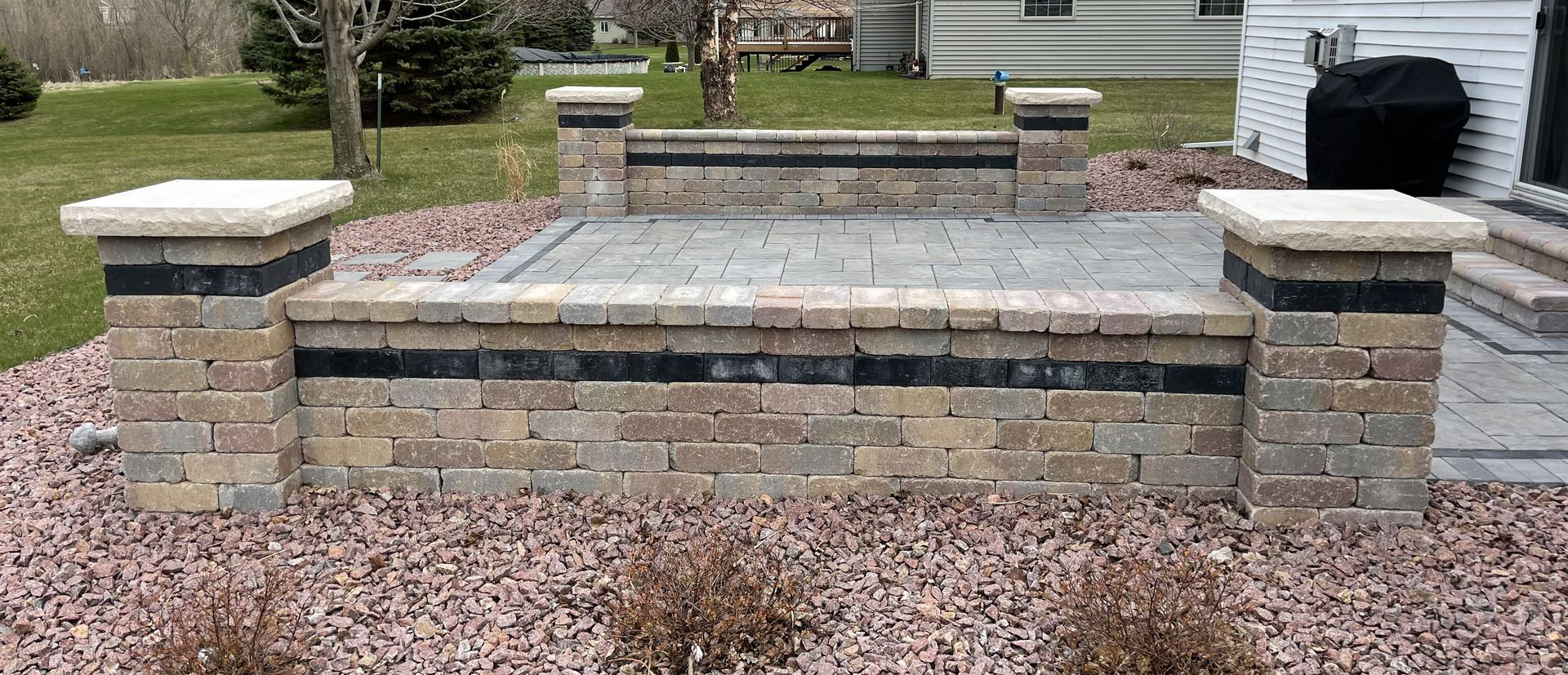 Stone backyard retaining wall with square pillars and a paved top, surrounded by mulch and grass.
