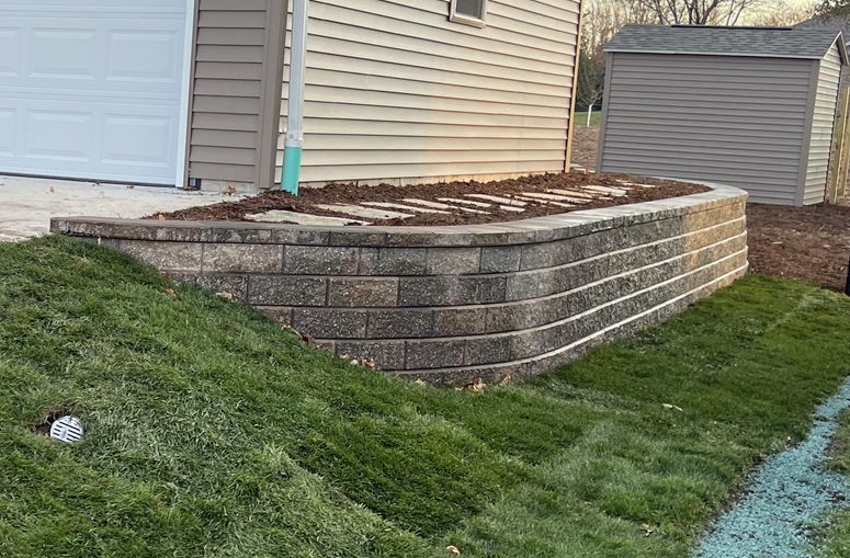 Retaining wall with stacked stone blocks beside a house, bordering a grassy sloped yard.