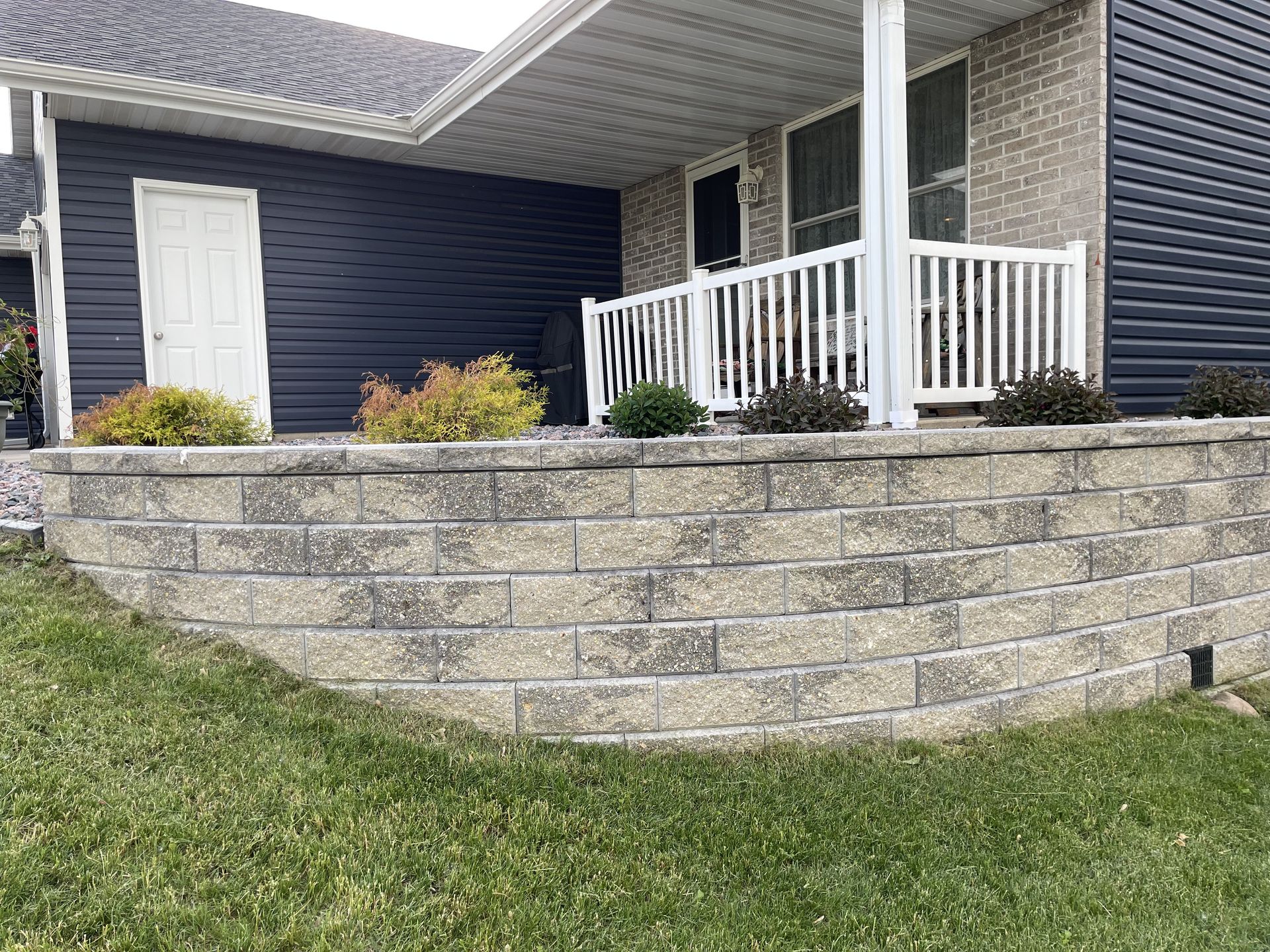 Stone retaining wall and front porch of a house with white railings and blue siding.