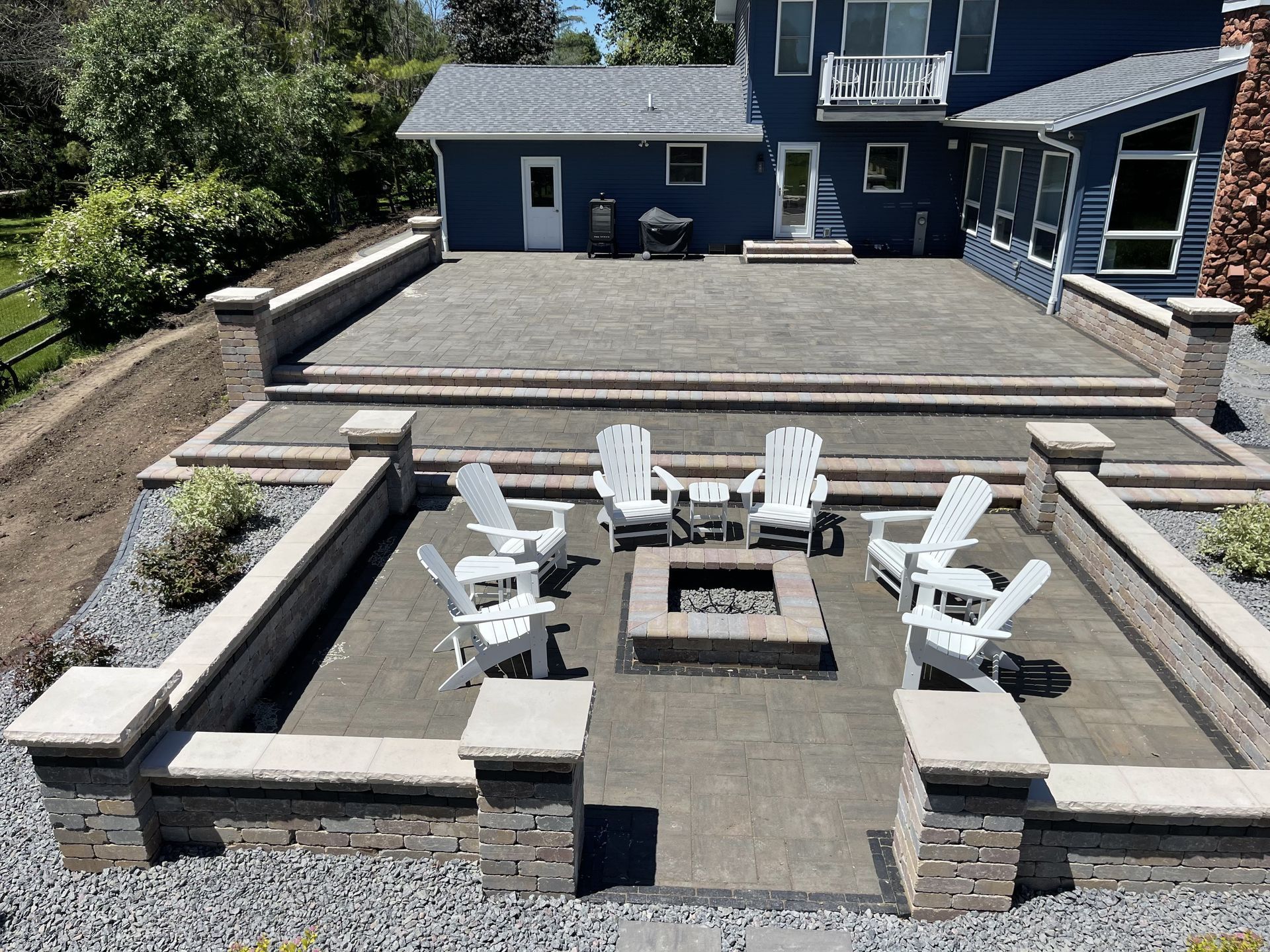 Backyard patio with stone fire pit and white chairs beside a blue house