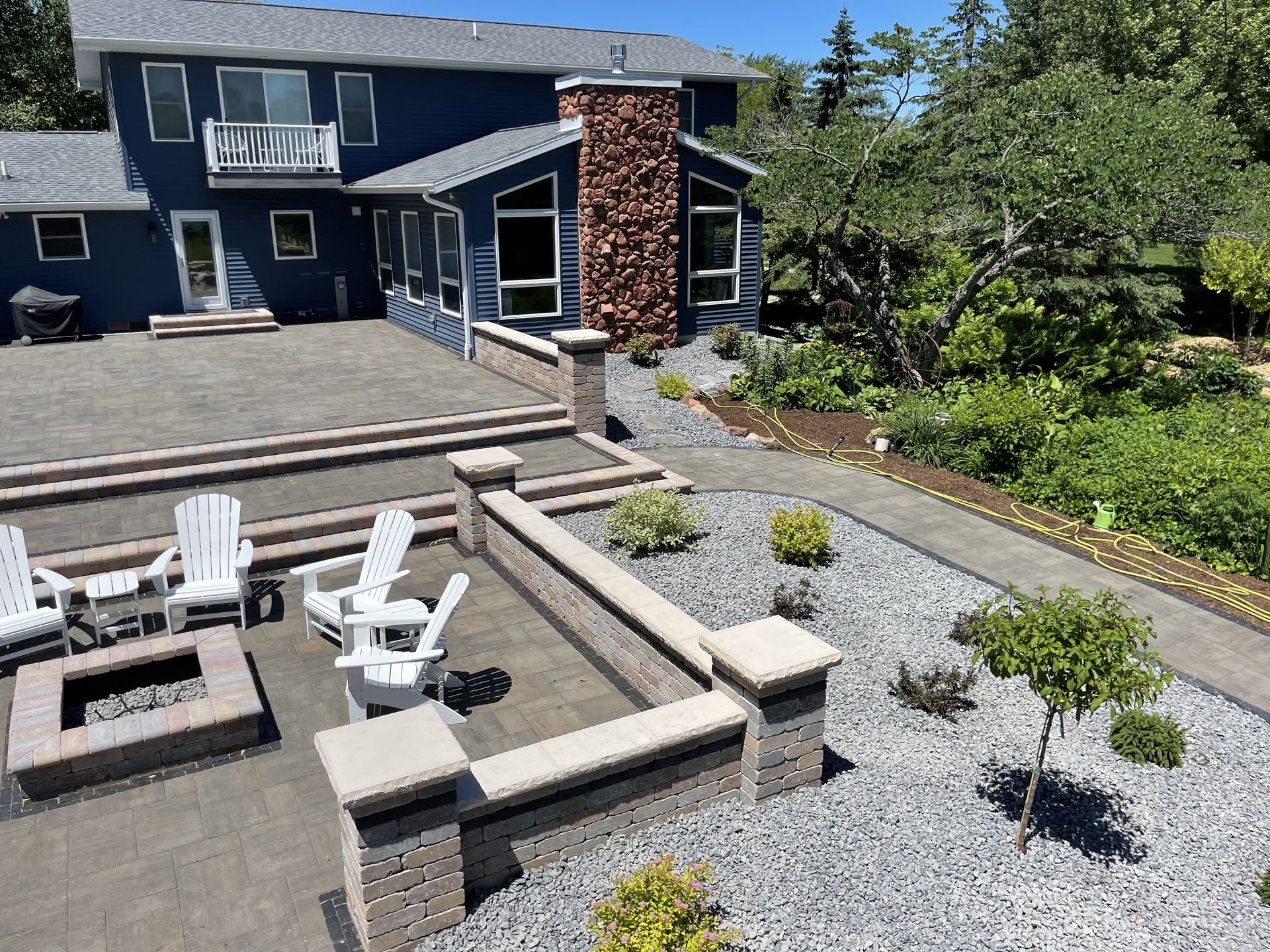 Backyard patio with fire pit, white chairs, stone path, and a blue house with brick chimney
