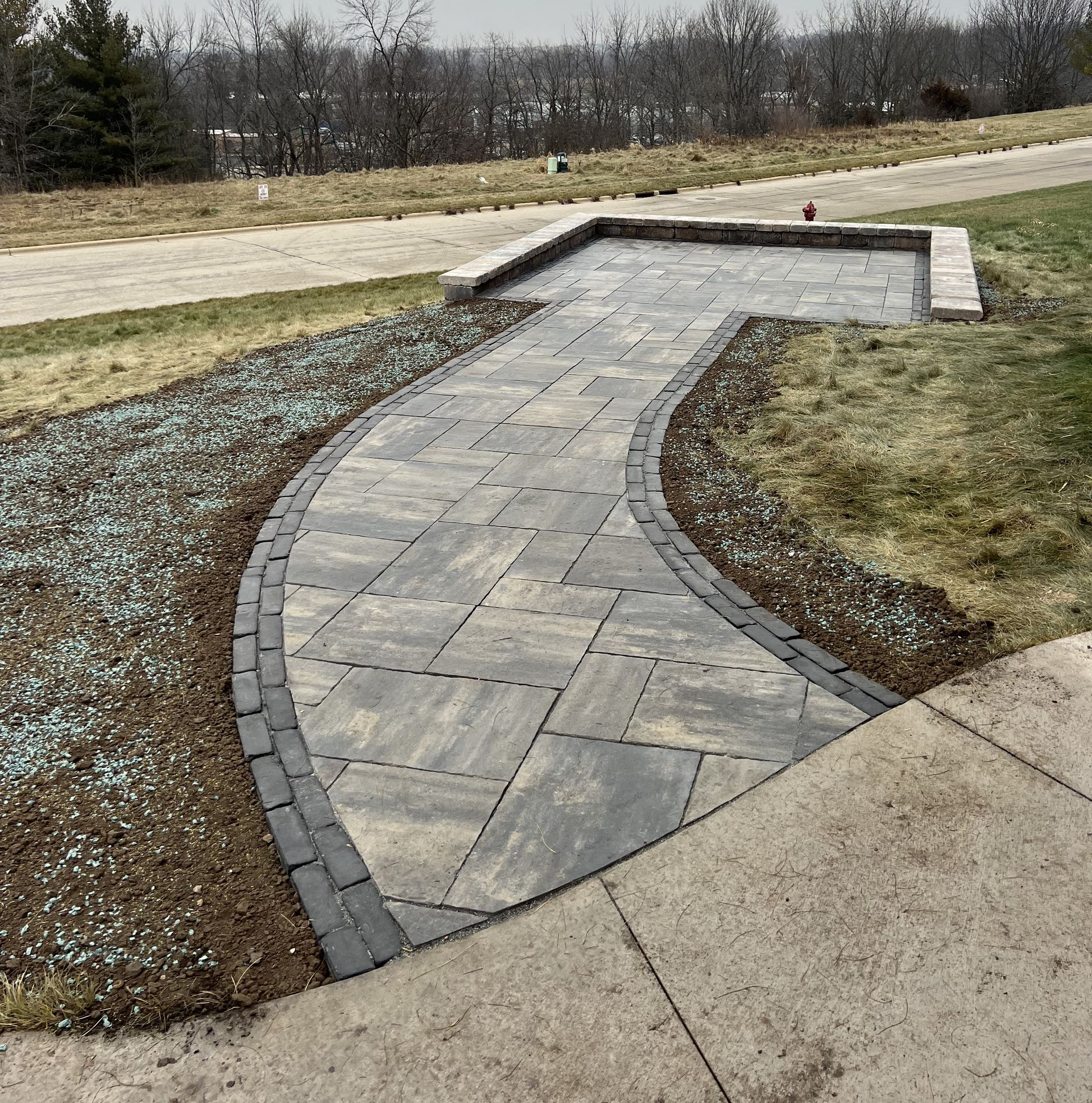 Curved stone walkway leading to a small bridge in a grassy park, bordered by gravel and concrete paths