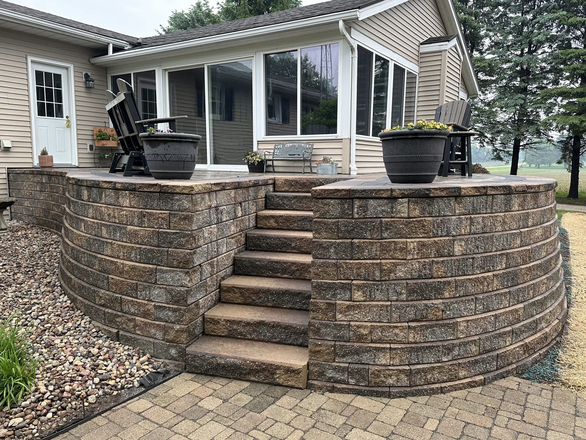 Stone patio with central steps leading to a house deck, flanked by potted planters.