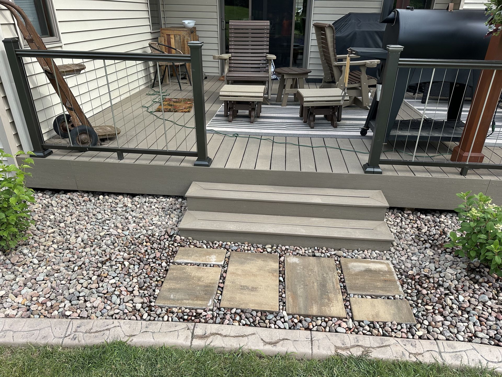 Backyard deck steps with railing, gravel border, and stone pavers leading to a patio.