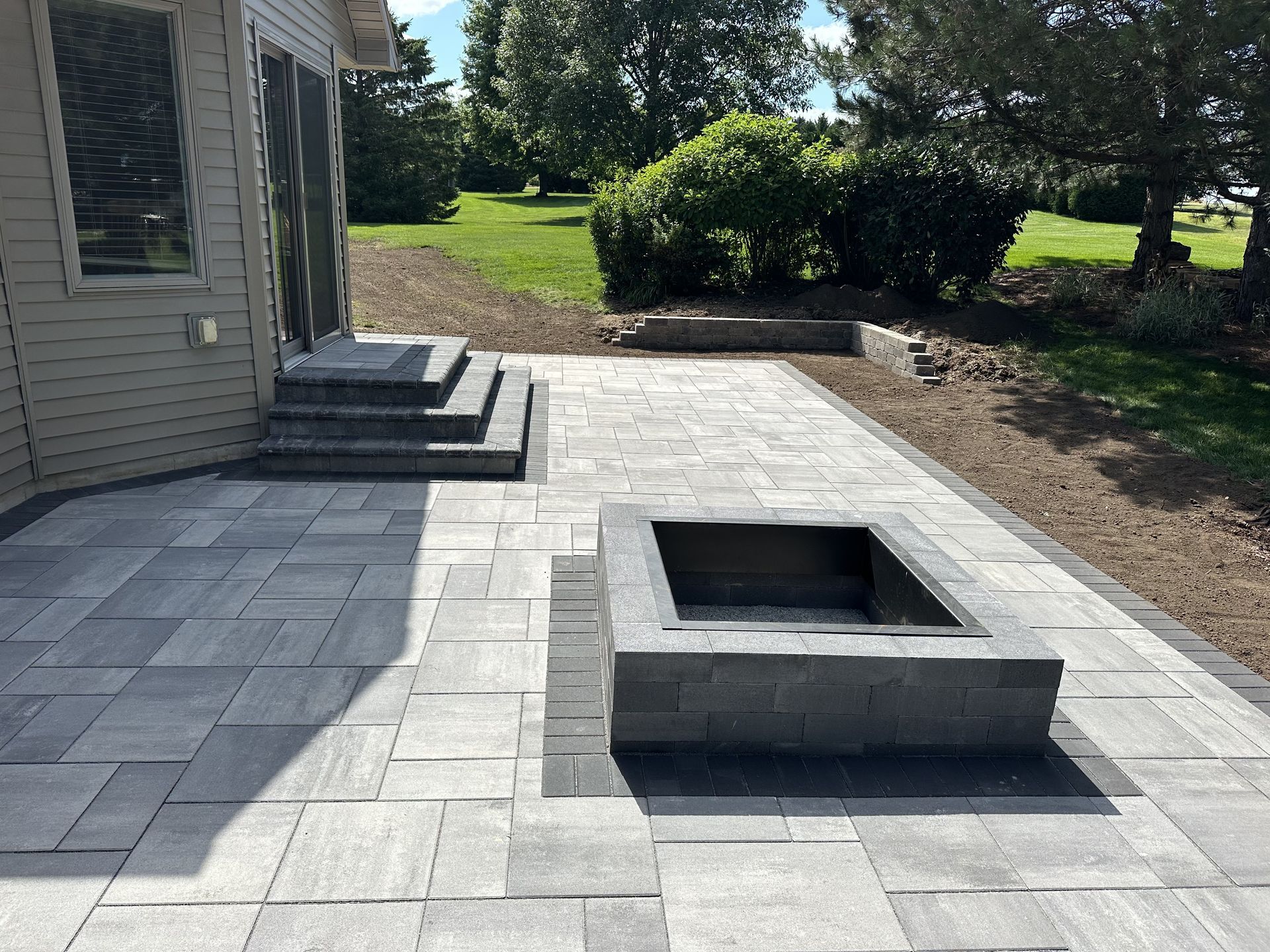 Backyard patio with stone pavers and a raised rectangular fire pit beside a house