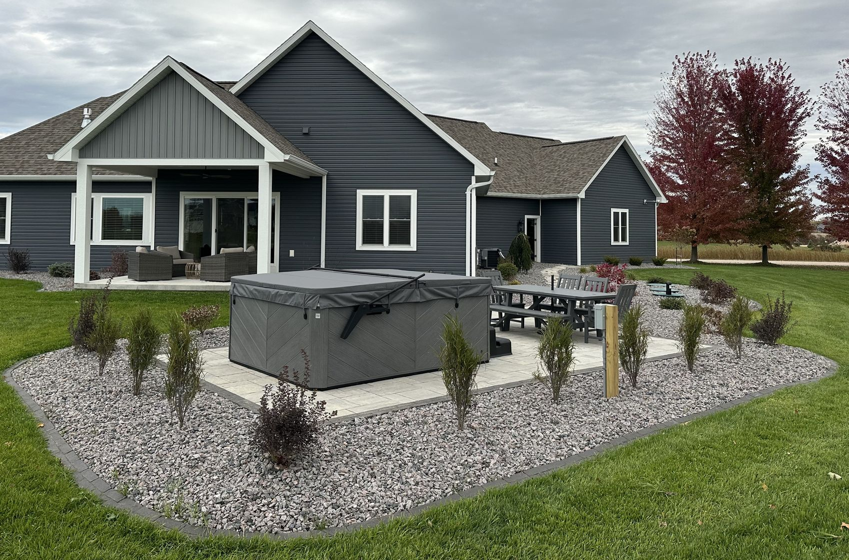 Modern dark-gray house with a gravel patio, hot tub, picnic table, and landscaped lawn under cloudy skies