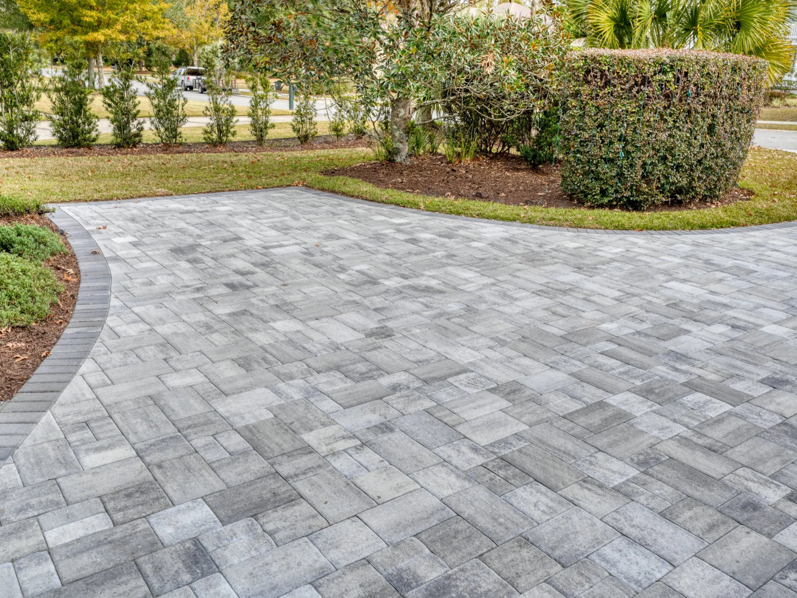 Gray paver driveway bordered by mulch, shrubs, and trees in a landscaped yard