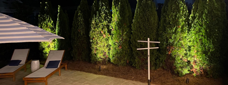 Nighttime backyard with lit green shrubs, lounge chairs, and a white signpost near a patio umbrella