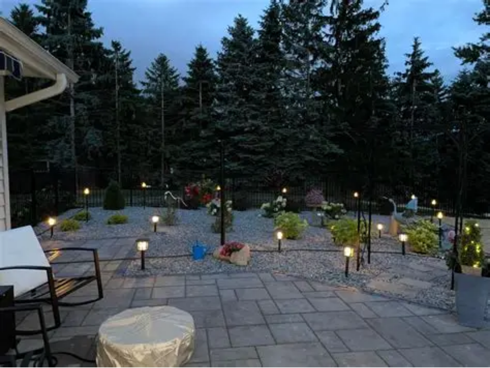 Stone patio at dusk with garden lights, chairs, and tall evergreens in the background.