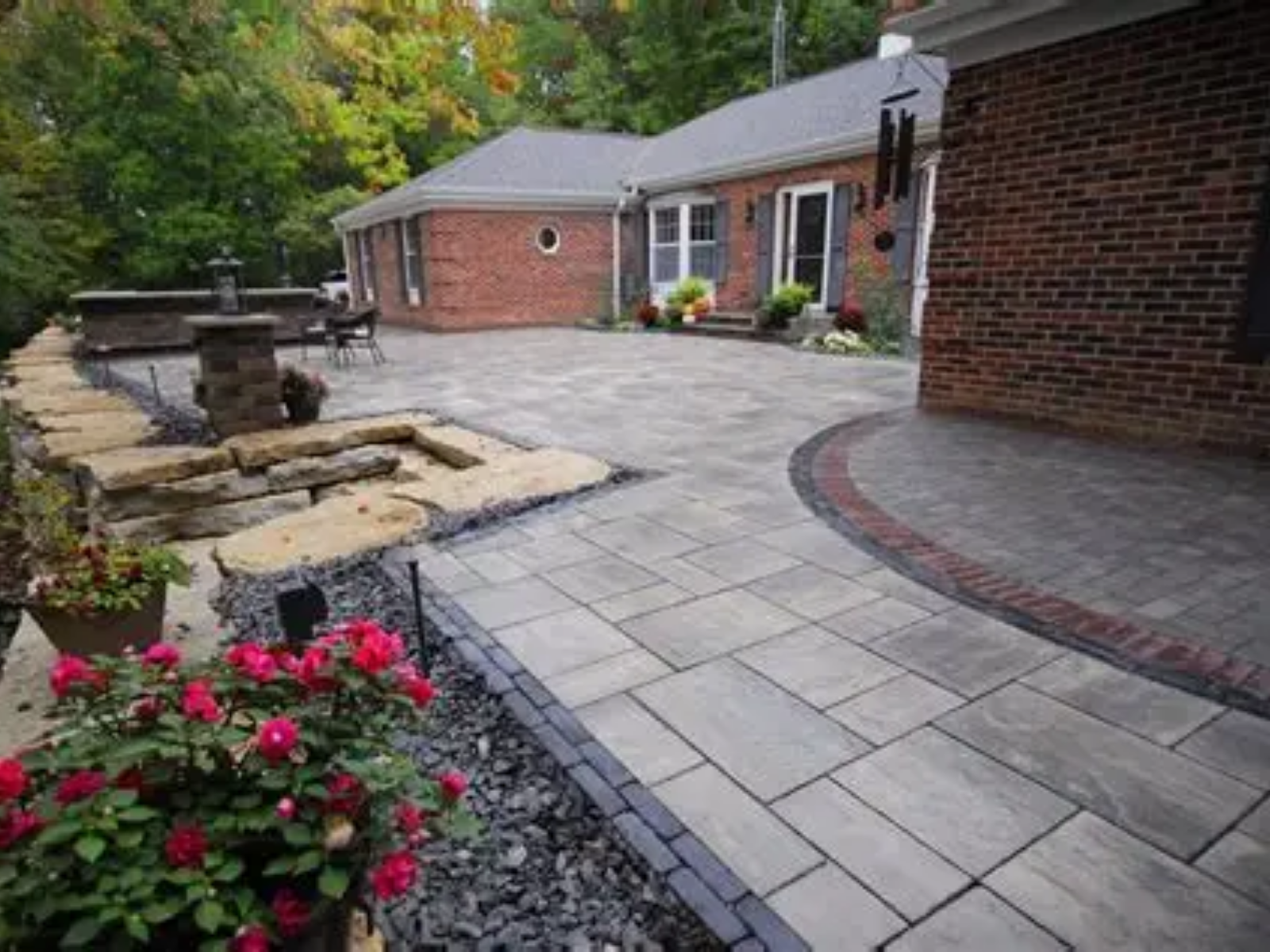 Brick patio with curved pavers, stone fountain, and pink flowers beside a brick house