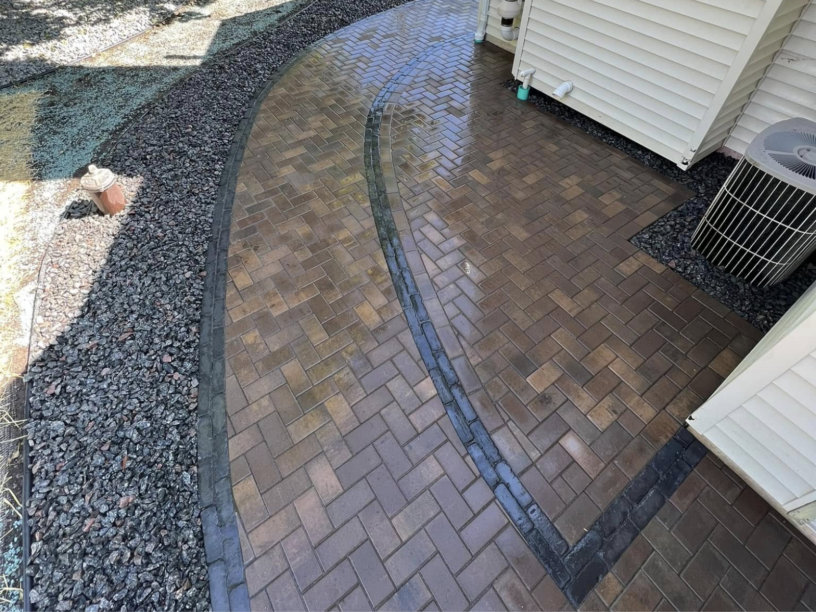 Curved brick patio and walkway beside a house, wet from rain, with gravel border and AC unit.